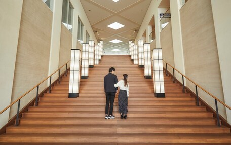 A staircase with wooden steps and black railings, flanked by walls with large windows and illuminated by modern light fixtures.