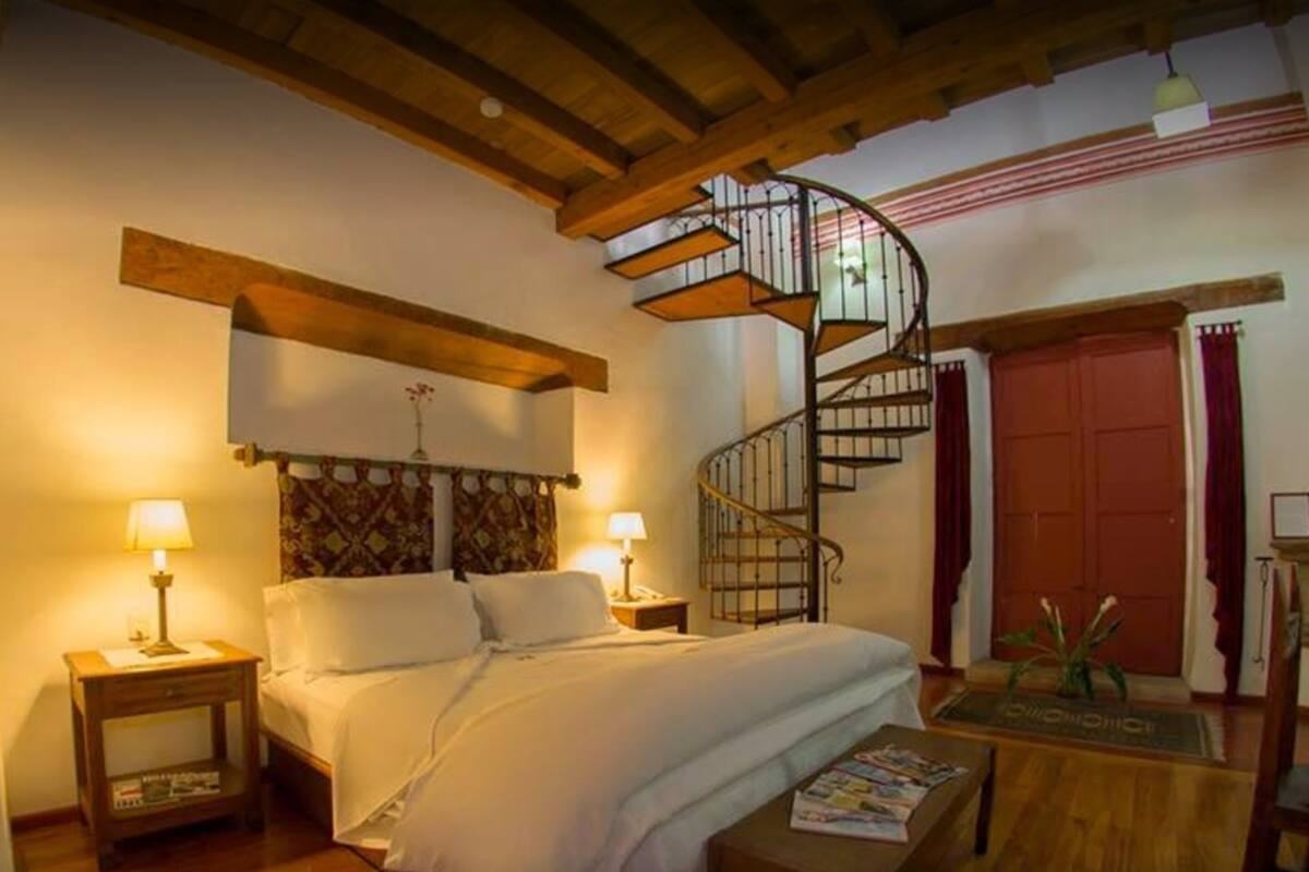 A bedroom with a wooden ceiling, a spiral staircase, a bed with white linens, two bedside tables with lamps, and a wooden floor.