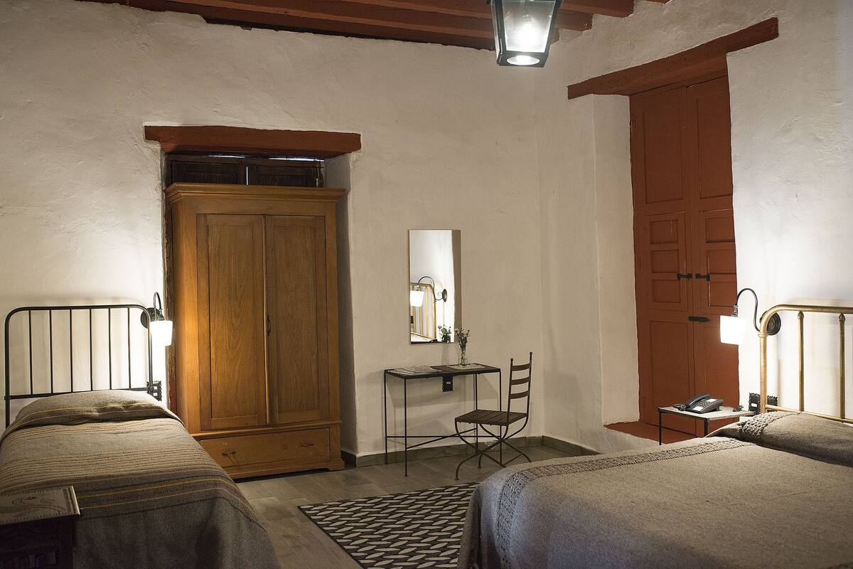 A bedroom with two beds, a wooden wardrobe, a desk with a chair, a mirror, and a door.