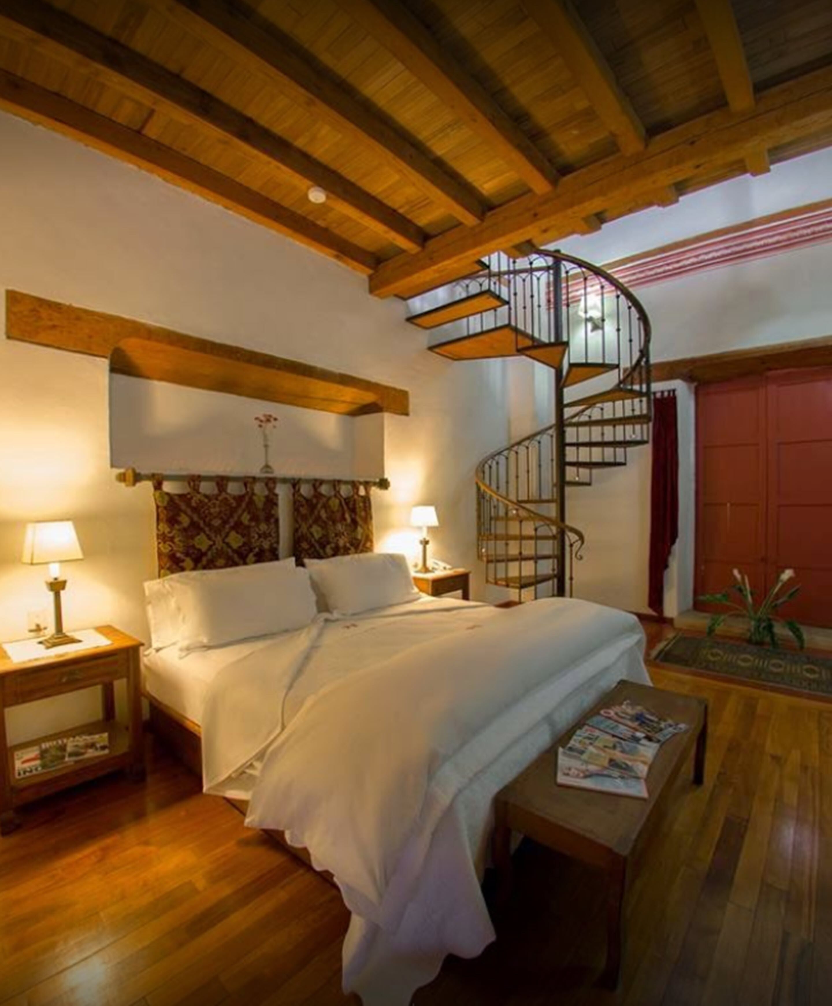 A bedroom with a wooden ceiling, a spiral staircase, a bed with white bedding, two bedside lamps, and a wooden floor.