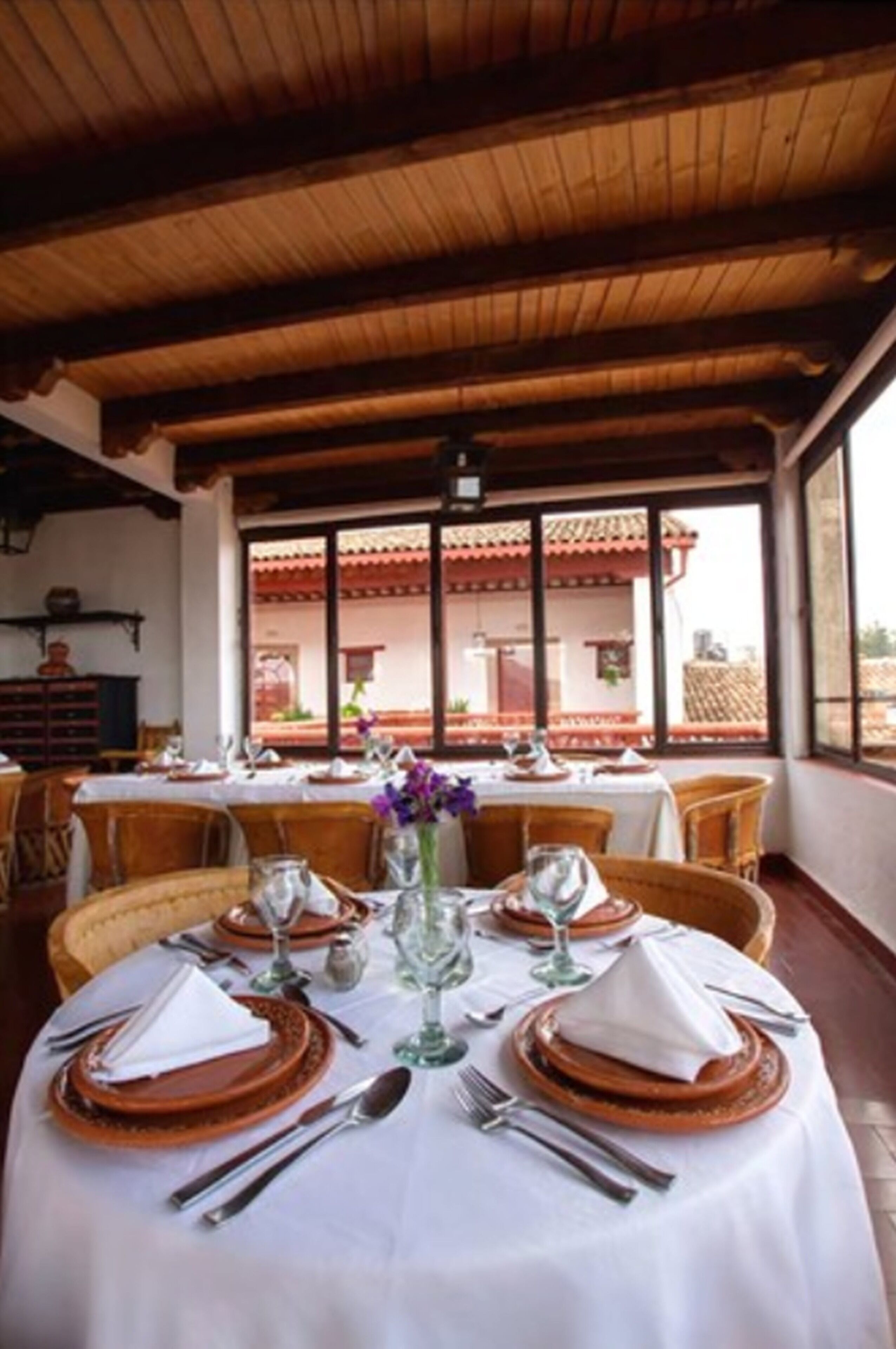 A dining area with round tables set for four, each with white tablecloths, brown plates, and silverware.