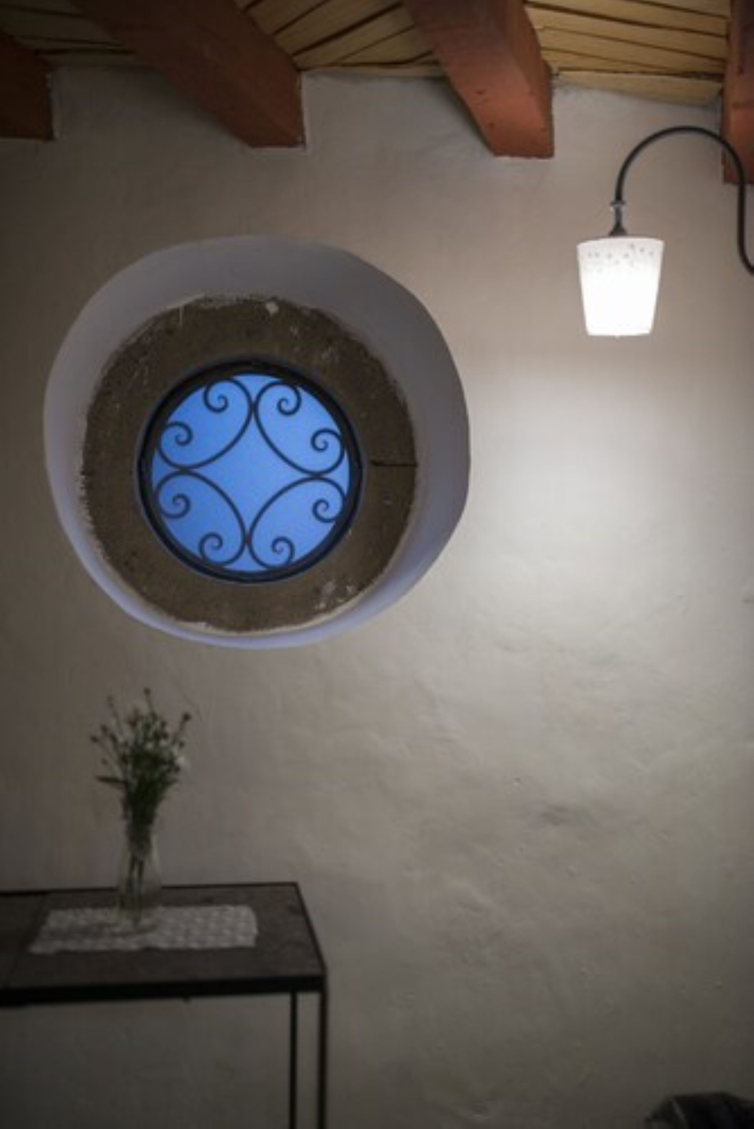 Una finestra circolare con un decorativo disegno in metallo, un punto luce a parete e un tavolino con un vaso.
