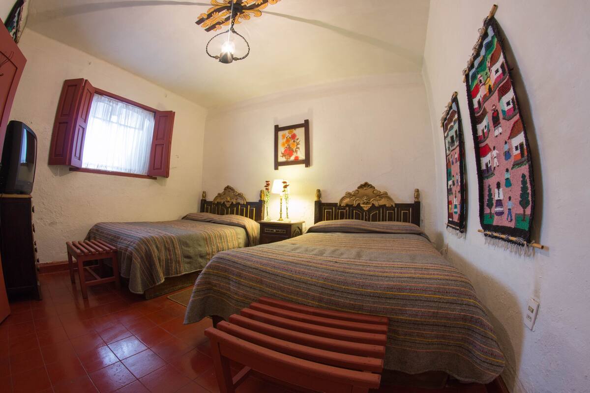 A bedroom with two beds, a wooden bench, a window with curtains, a bedside table with a lamp, and a wall hanging.