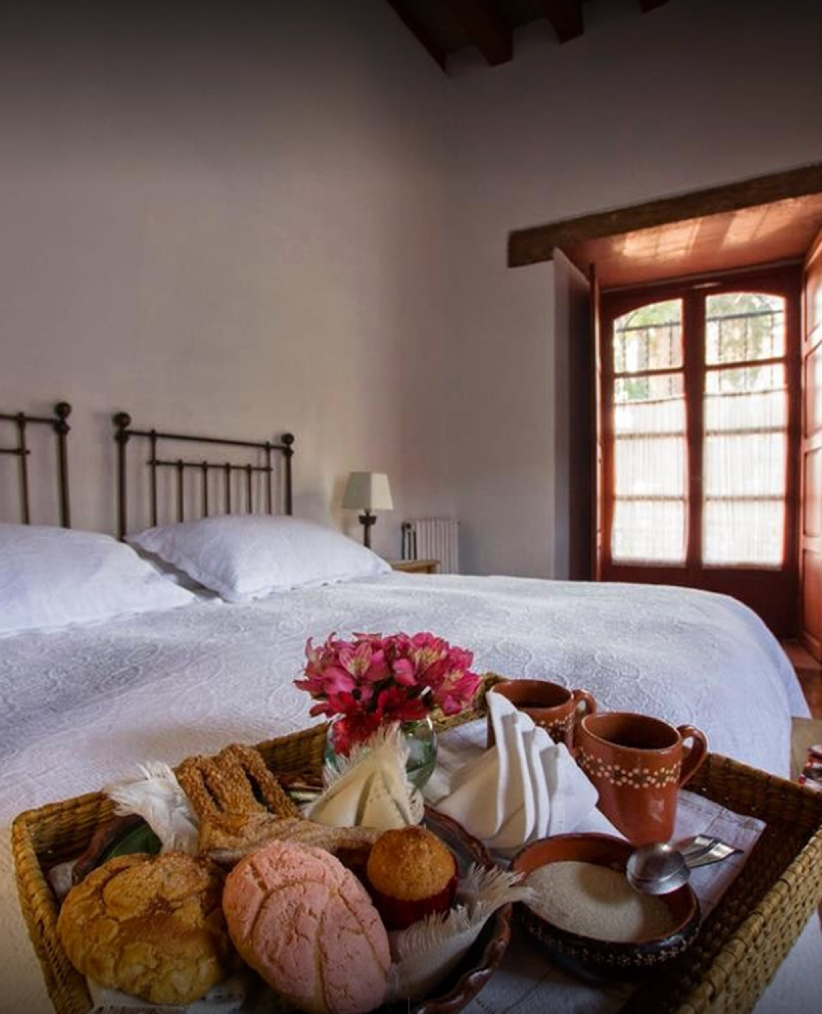 A neatly made bed with white linens, a basket of bread and pastries, a vase with flowers, and a cup and saucer on a tray.