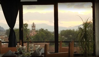 Mesa de jantar posta com copos e um vaso, com vista para uma paisagem montanhosa e um edifício ao longe.