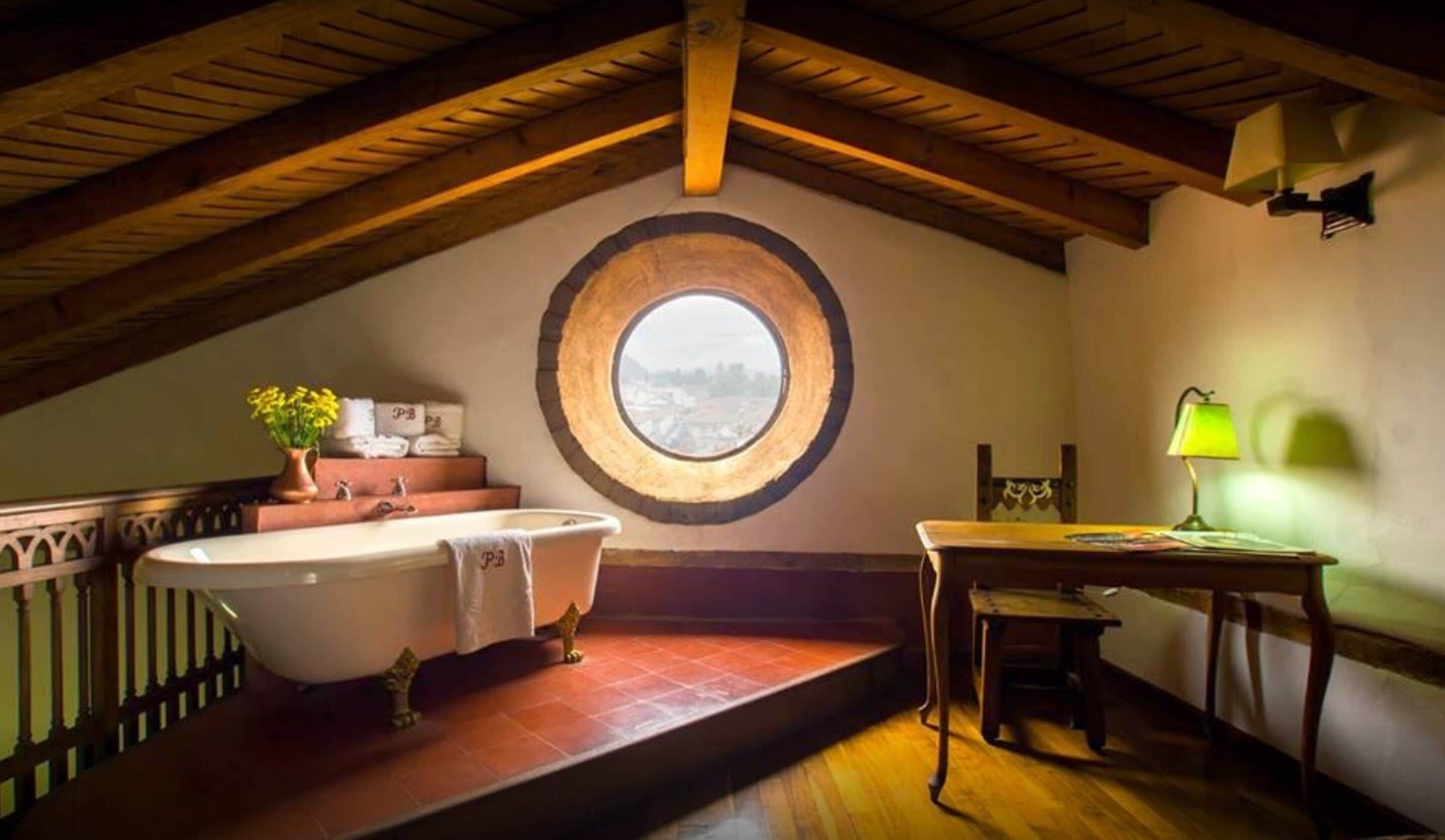 Un baño con una bañera independiente, un ventanal redondo, una mesa de madera y una silla.