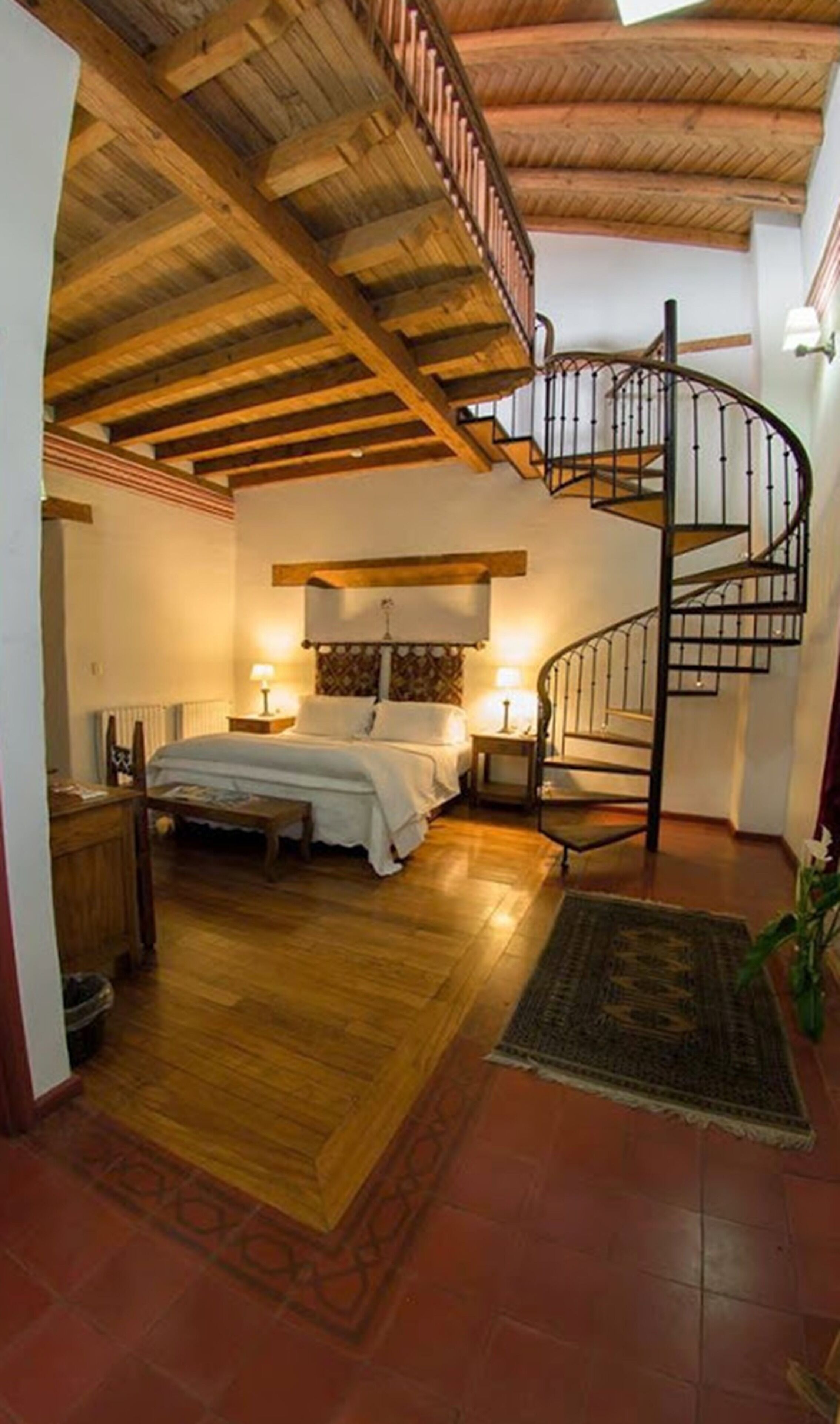 A bedroom with a wooden ceiling, a spiral staircase, a bed with a nightstand, and a radiator.