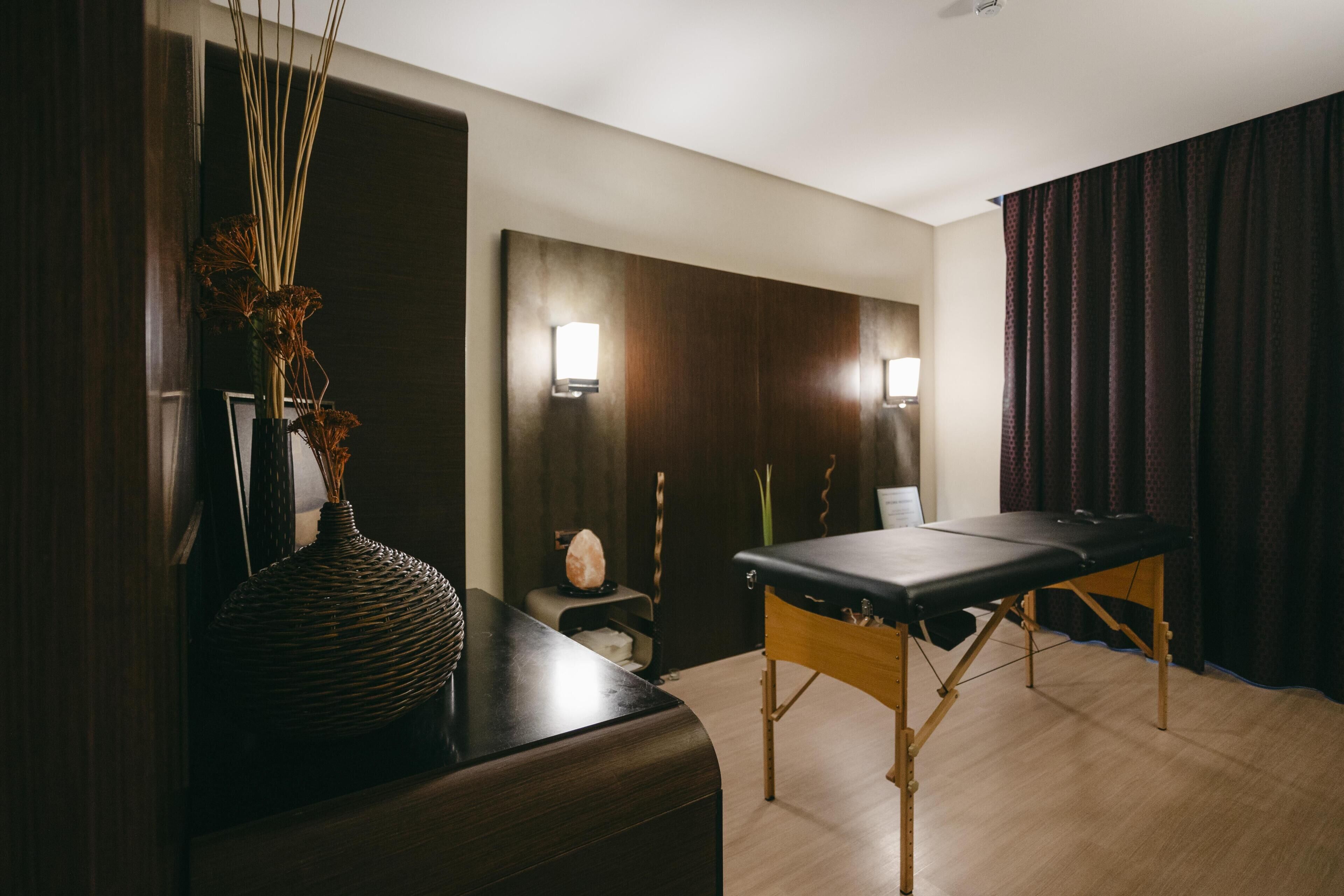 A massage table, a dark wooden cabinet, and a vase with dried plants.