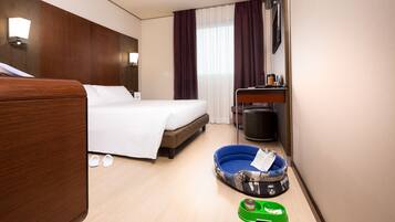 A hotel room with a bed, a desk, a chair, a pet bed, and a trash bin.
