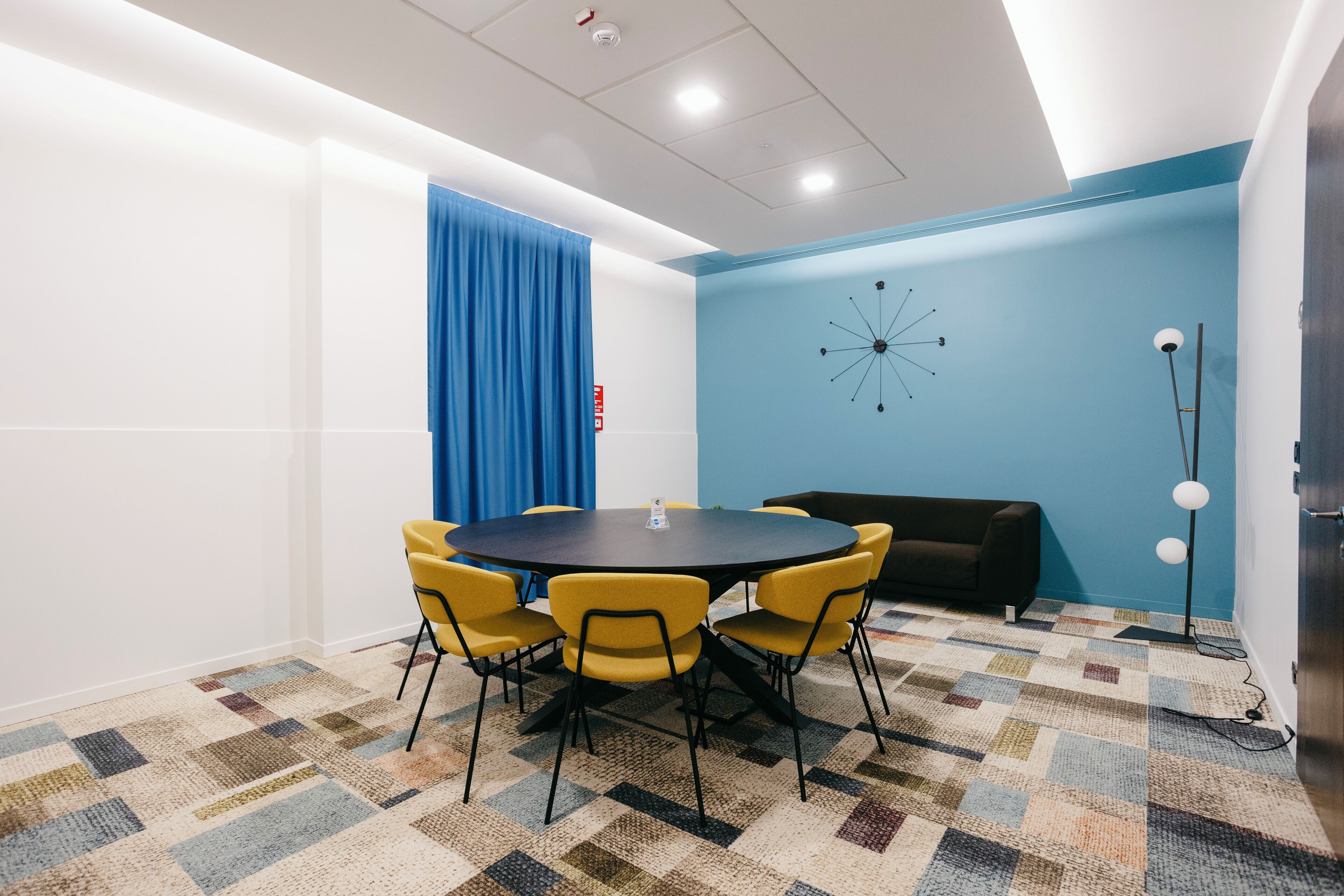 A conference room with a round table, yellow chairs, a blue curtain, a black sofa, a patterned carpet, and a wall clock.