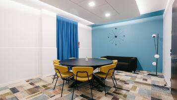 A conference room with a round table, yellow chairs, a blue curtain, a black sofa, a patterned carpet, and a wall clock.