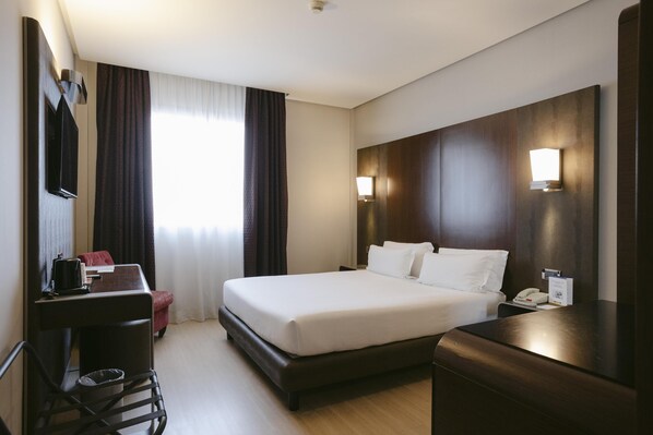 Ein Hotelzimmer mit einem großen Bett, einem Schreibtisch mit Stuhl, einem kleinen Tisch, einem Telefon und einer Lampe.