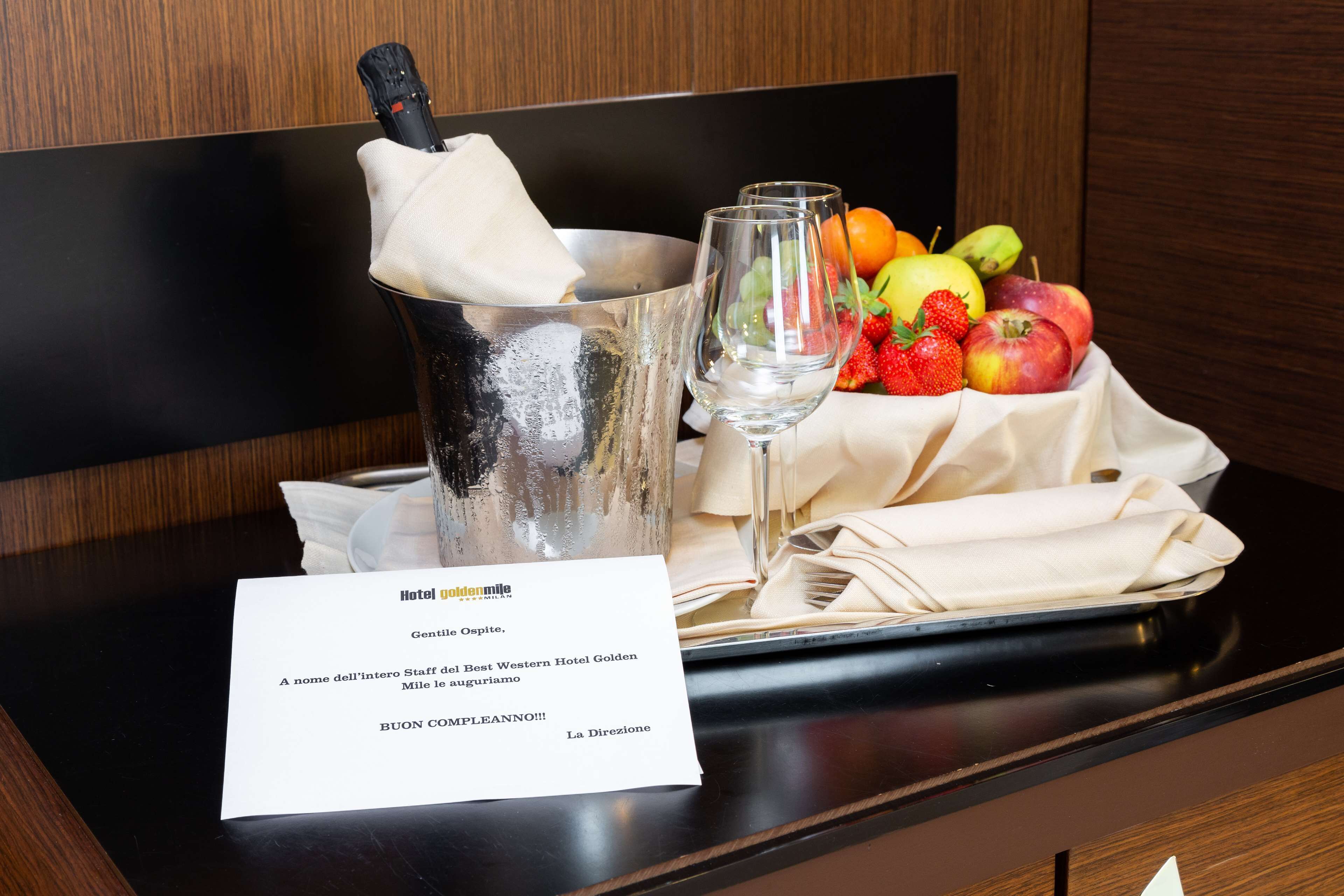 A hotel room with a minibar containing a bottle of champagne, a glass, and a fruit basket. A note from the hotel staff is also visible.