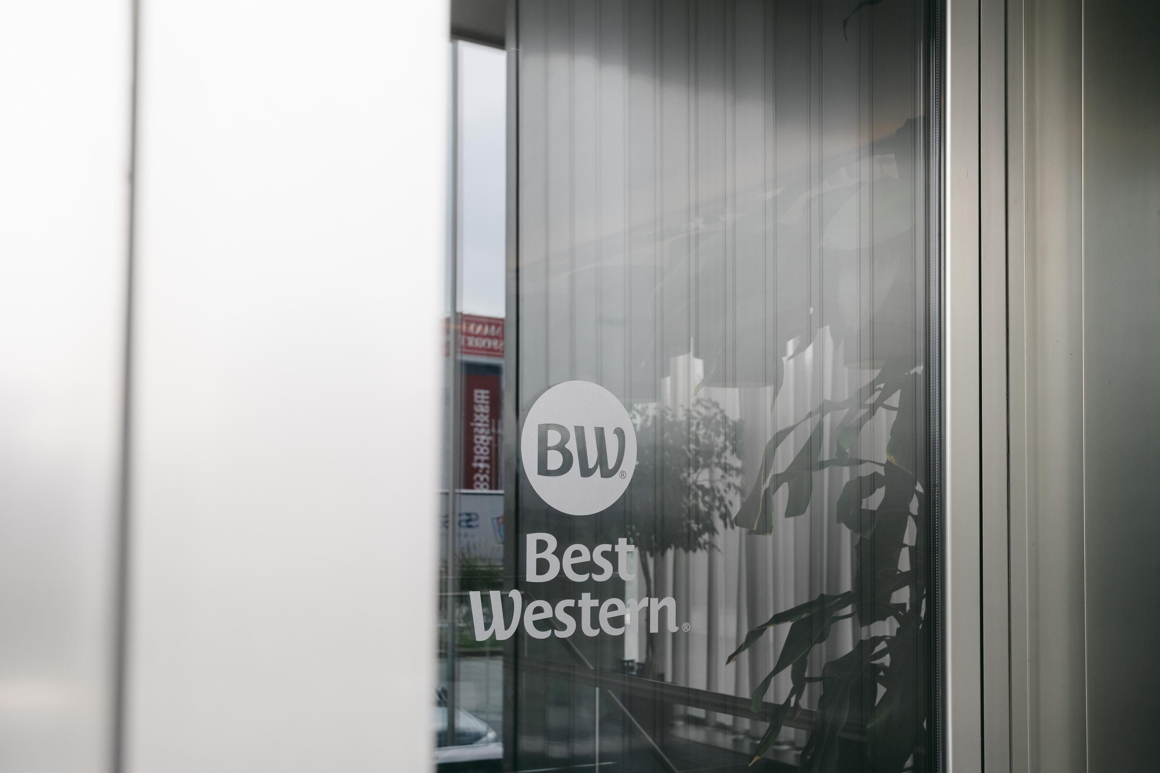 Una ventana de vidrio con el logotipo 'BW Best Western' y el reflejo de un edificio y algunas plantas.