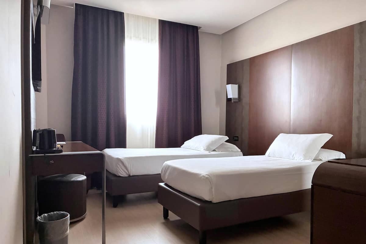 Une chambre d’hôtel avec deux lits, un bureau, une poubelle et une fenêtre avec des rideaux.