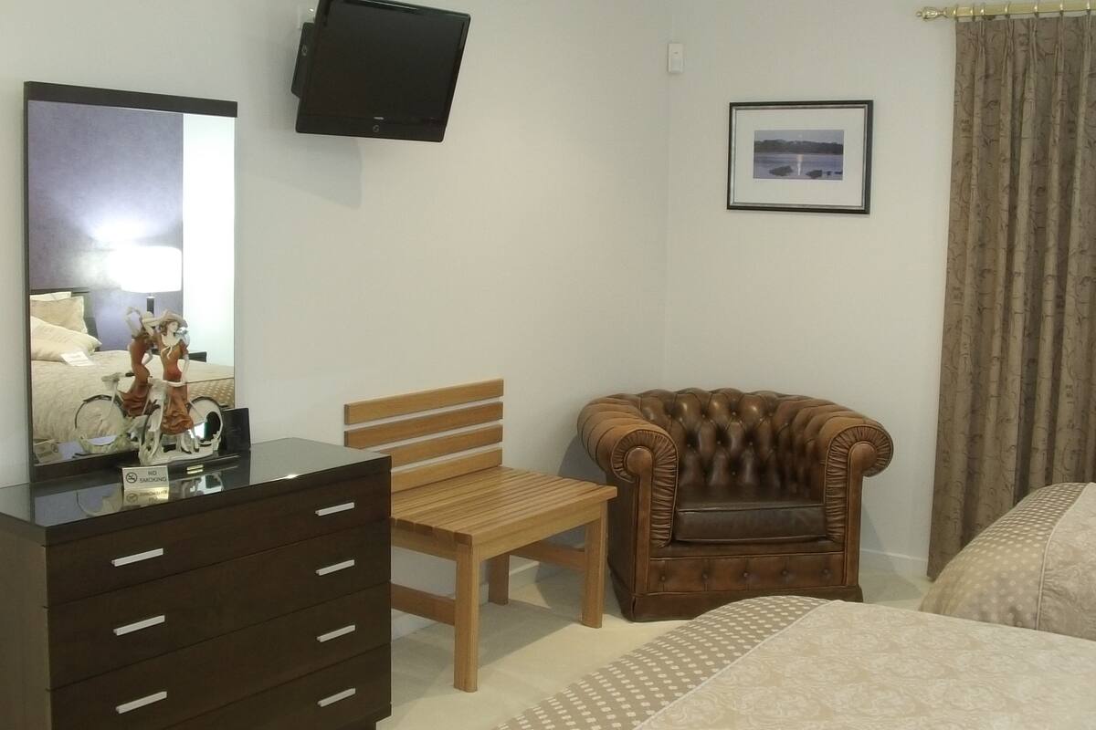 A hotel room with a wooden dresser, a brown leather armchair, a wooden chair, a bed, a television mounted on the wall, and a framed picture on the wall.