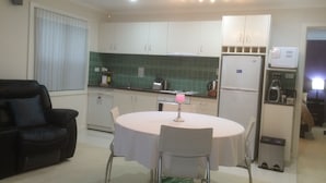 Een kleine keuken met witte kastjes, een eettafel met stoelen, een zwarte leren bank en een kleine prullenbak.