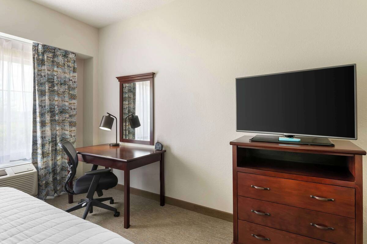 A hotel room with a bed, a desk with a chair, a flat-screen TV on a wooden stand, and a mirror above the desk.