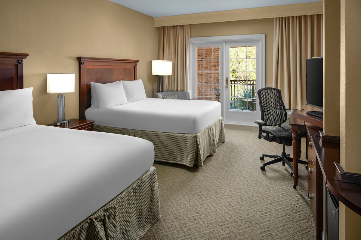 A hotel room with two beds, a desk with a chair, a television, and a balcony with a view.