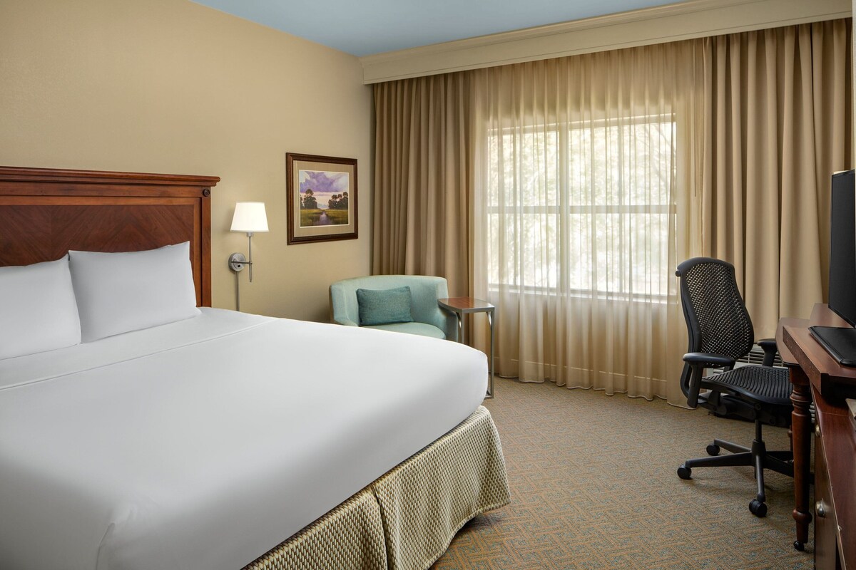 A hotel room with a large bed, a desk with a computer, a chair, and a window with curtains.