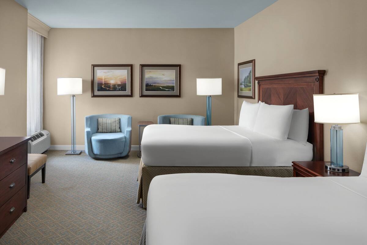 A hotel room with two beds, a desk, a chair, and two lamps.
