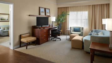 A hotel room with a desk, chair, television, sofa, and ottoman.