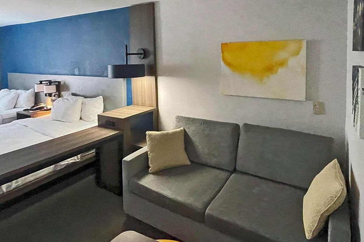 A modern hotel room with a bed, a grey sofa, a small table, and a wall-mounted lamp.