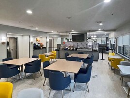 A modern dining area with tables and chairs, a kitchen in the background, and a view of the city through large windows.
