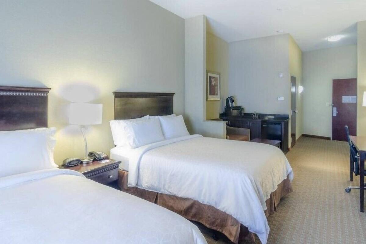A hotel room with two beds, a desk, a chair, a lamp, and a small kitchenette.