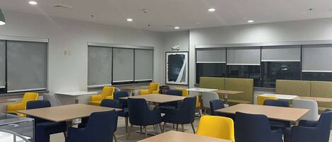 A modern classroom with tables and chairs, large windows, and a projector screen.