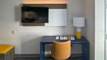 Una habitación de hotel con escritorio, silla, televisor, lámpara y una mesita con objetos encima.