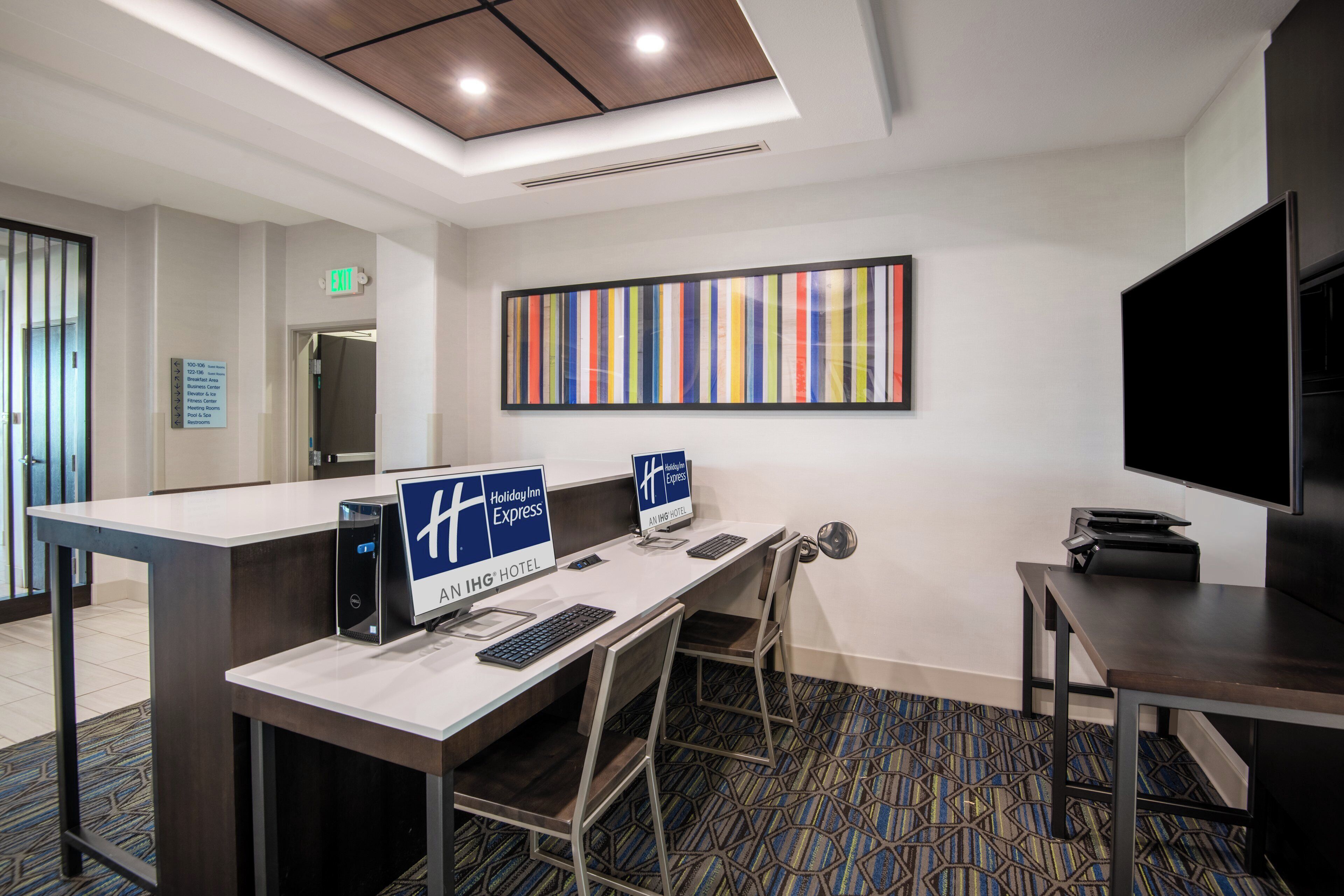 A hotel reception area with a desk, computer, and chairs. There is a large framed artwork on the wall and a television on a stand.