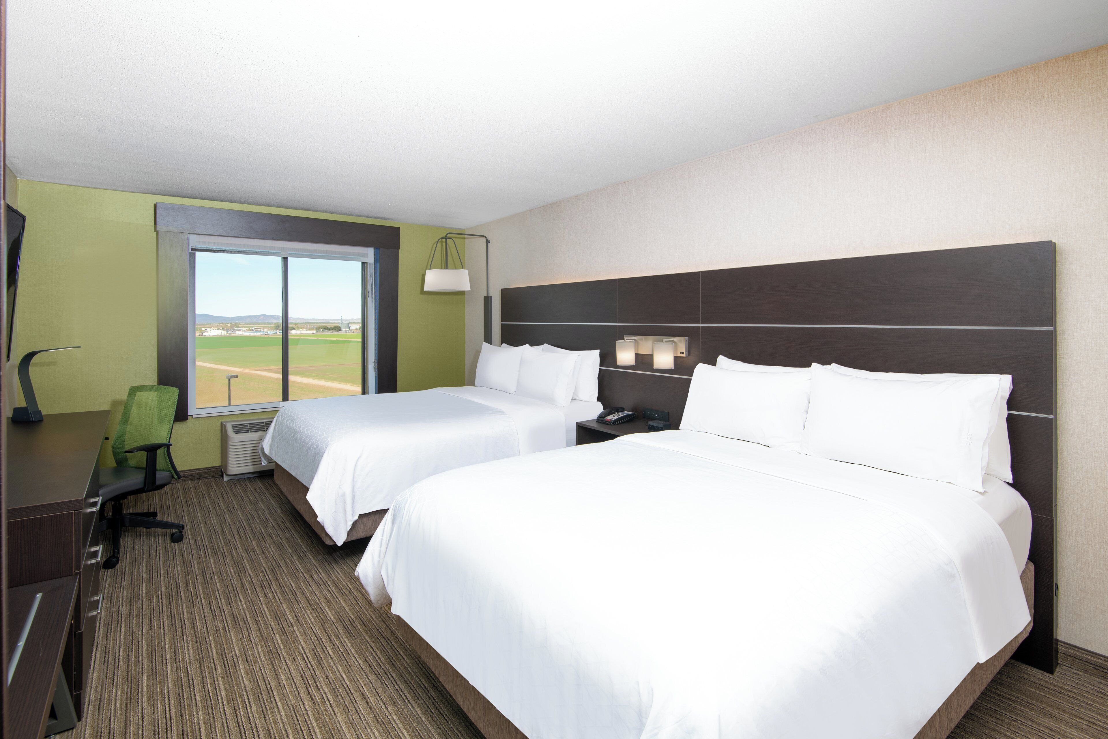 A hotel room with two beds, a desk, a chair, and a large window offering a view of a field.