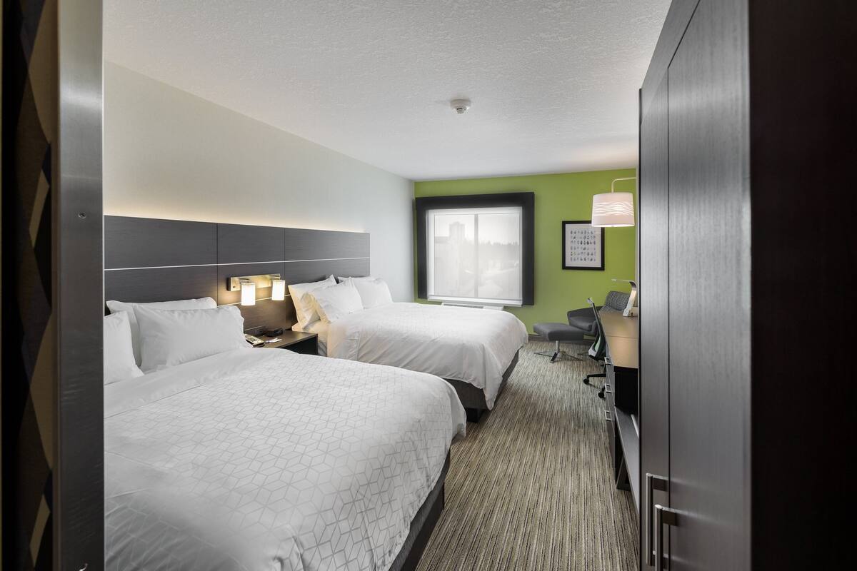 A hotel room with two beds, a desk, a chair, a window with a city view, and a green accent wall.