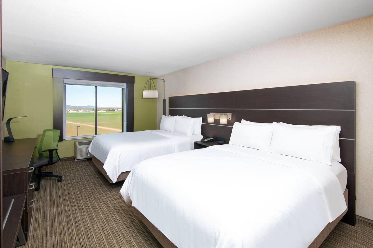 A hotel room with two beds, a desk, a chair, a lamp, and a large window with a view of a baseball field.