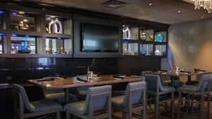 A bar area with multiple tables and chairs, shelves with decorative items, and a flat-screen TV mounted on the wall. - The Rewind West Des Moines, Tapestry Collection by Hilton (West Des Moines)