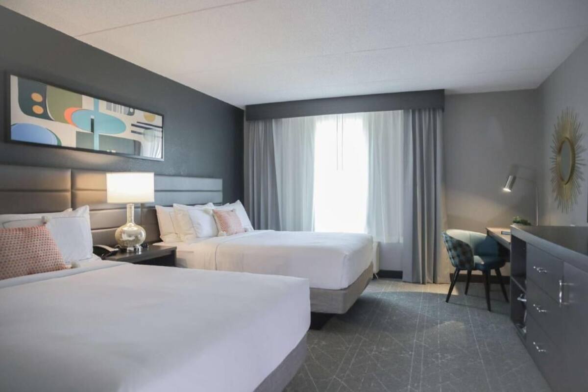 A hotel room with two beds, a desk, a chair, a lamp, and a large window with curtains.
