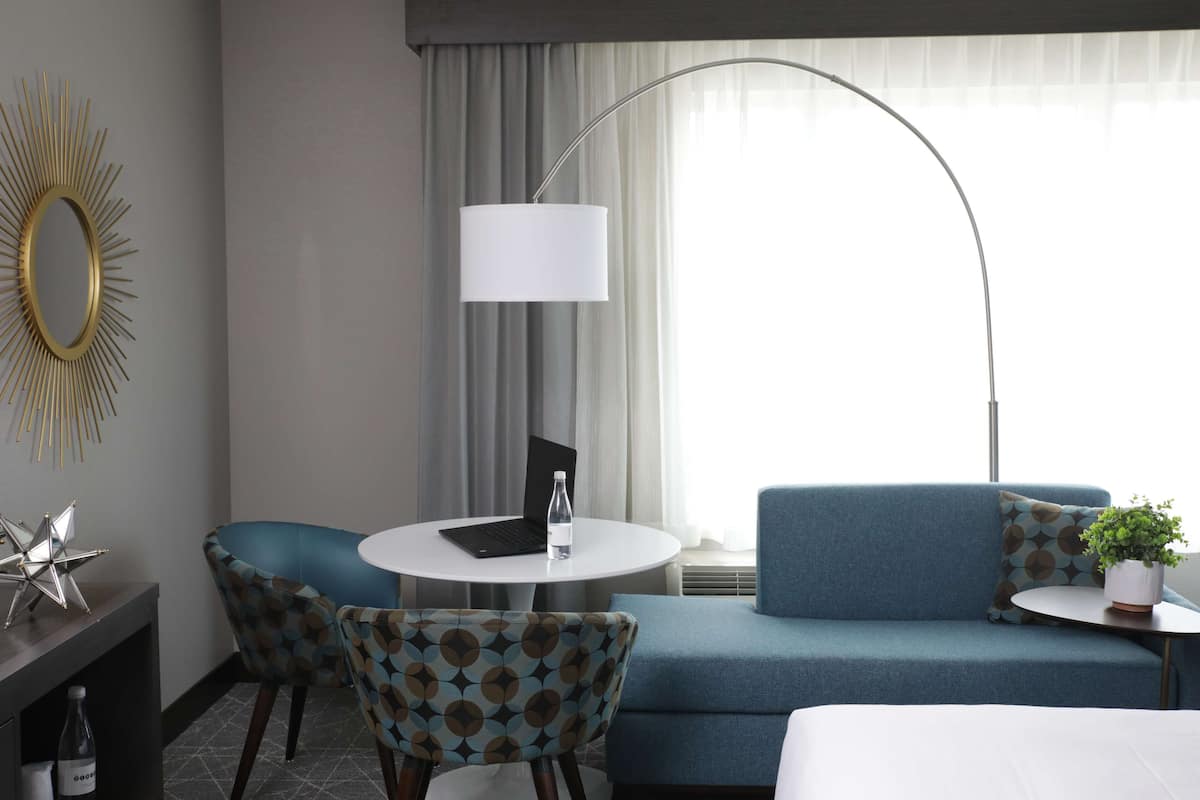 A modern hotel room with a blue sofa, a round white table, a laptop, a lamp, and a decorative mirror.