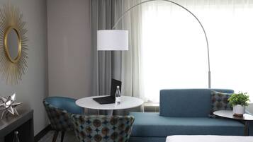 Une chambre d’hôtel moderne avec un canapé bleu, une table ronde blanche, un ordinateur portable, une lampe et un miroir décoratif.