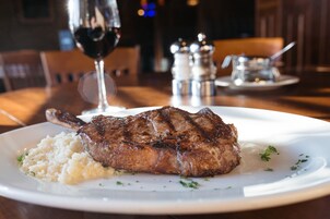 A steak with a side of rice on a white plate, set on a wooden table with a glass of red wine and salt and pepper shakers in the background.