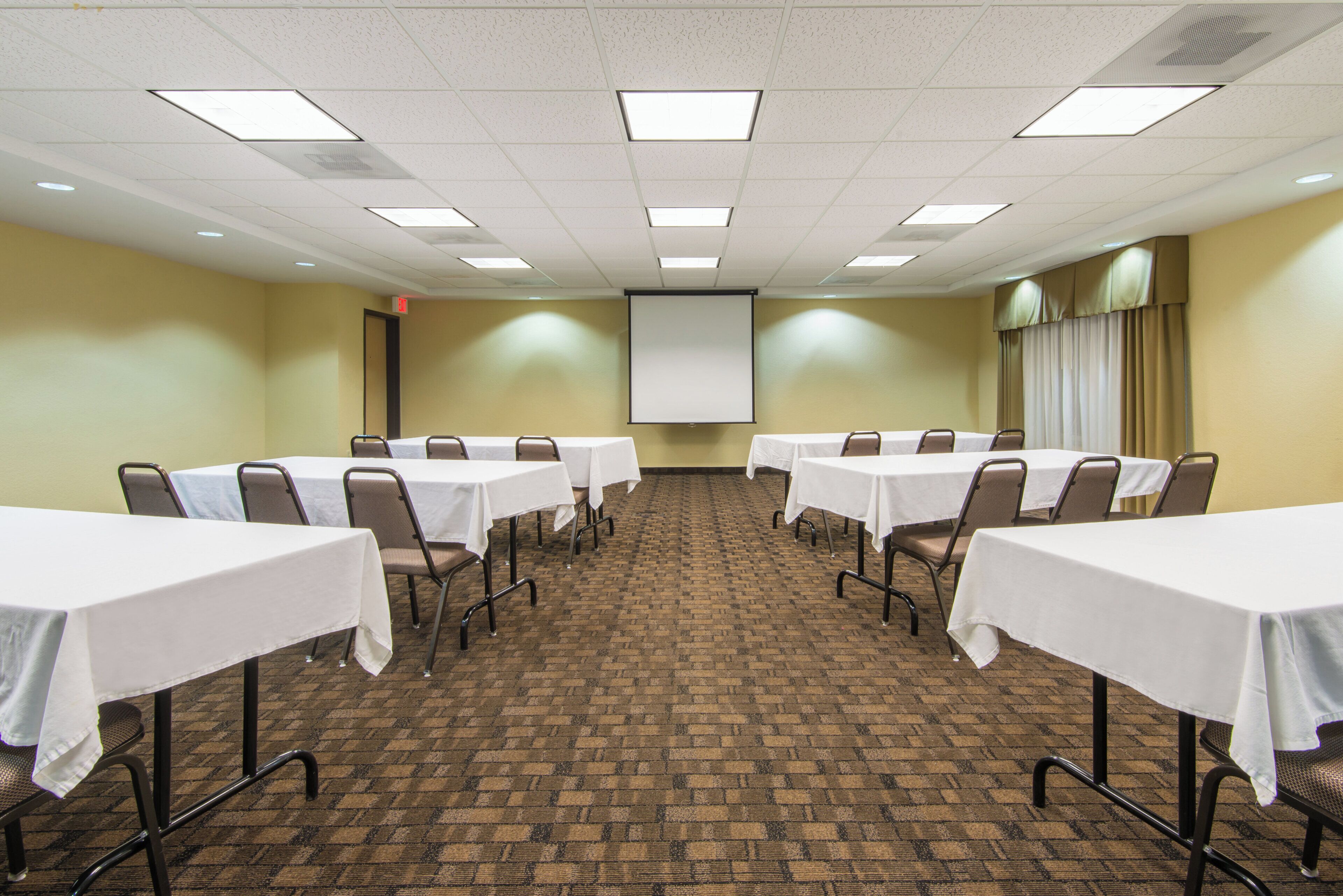 A conference room with multiple tables and chairs, a projector screen, and a carpeted floor.