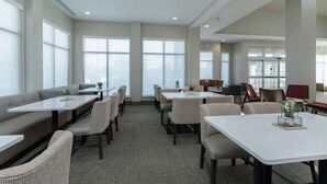 A spacious indoor area with multiple seating arrangements including tables and sofas, large windows providing natural light, and a clean, modern design.