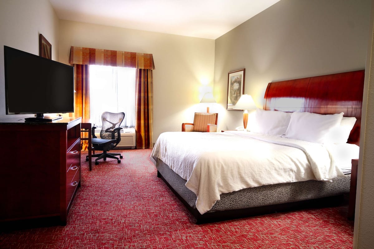 Ein Hotelzimmer mit Bett, Schreibtisch mit Fernseher, Sessel, Lampe und einem Bild an der Wand.
