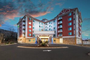 Un hotel de varios pisos con entrada central, estacionamiento al frente y montañas al fondo.