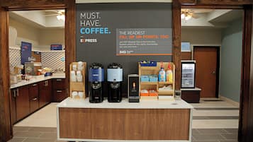 Una estación de café con un cartel que promociona el Club Rewards IHG, una cafetera, un dispensador de agua caliente y un refrigerador con bebidas.
