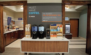 Una estación de café con un cartel que promociona el Club Rewards IHG, una cafetera, un dispensador de agua caliente y un refrigerador con bebidas.