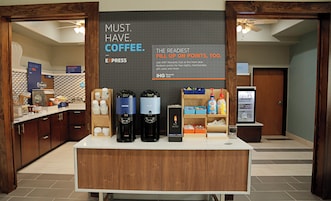Una estación de café con un cartel que promociona el Club Rewards IHG, una cafetera, un dispensador de agua caliente y un refrigerador con bebidas.
