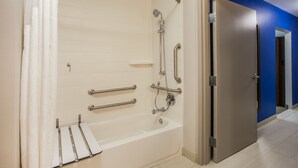 Een badkamer met een badkuip, handgrepen, een douchekop en een deur naar een andere kamer.