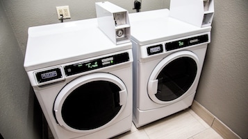 Two white front-loading washing machines with digital displays, placed next to each other in a corner with a light-colored tiled floor.
