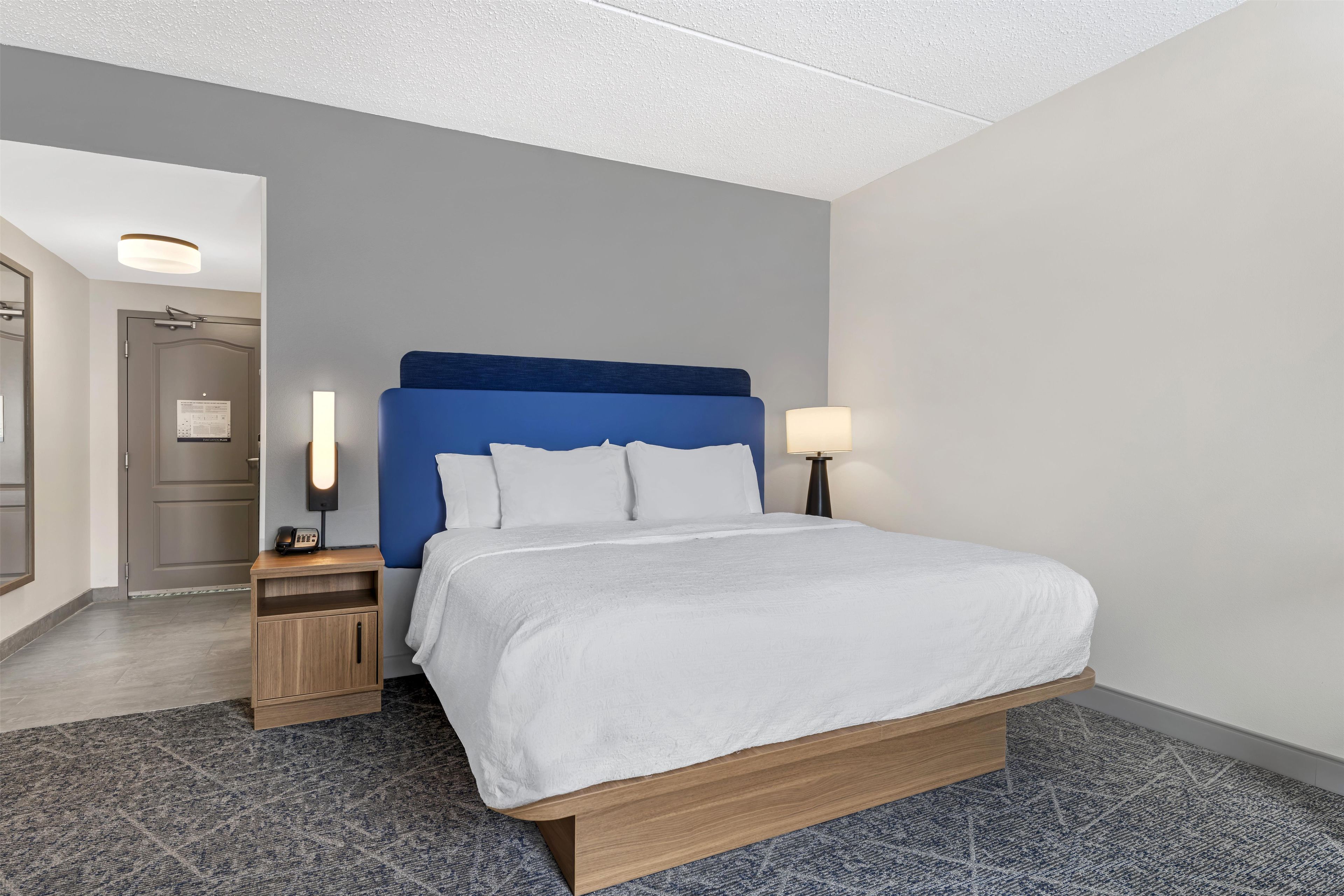 Une chambre d’hôtel avec un lit, des tables de chevet, une commode, une lampe et une porte.